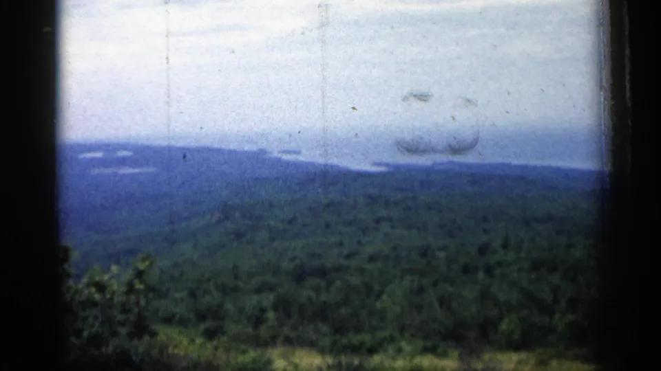 1960 Michigan home-movie still of rolling green woodland and distant blue water, framed by dark film or window edges.