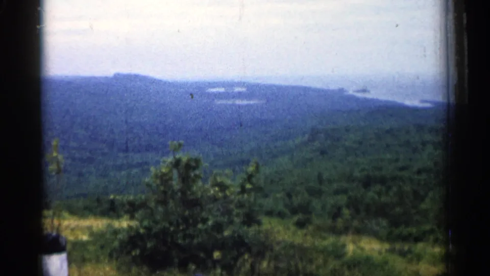 1960 Michigan home-movie frame showing foreground shrubs, rolling forest, blue hills, and distant Great Lakes water.