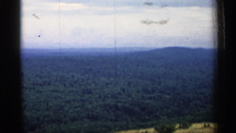 1960 Michigan 8mm frame of a wooded ridge and cloudy sky, with soft focus, dust marks, and edge darkening.