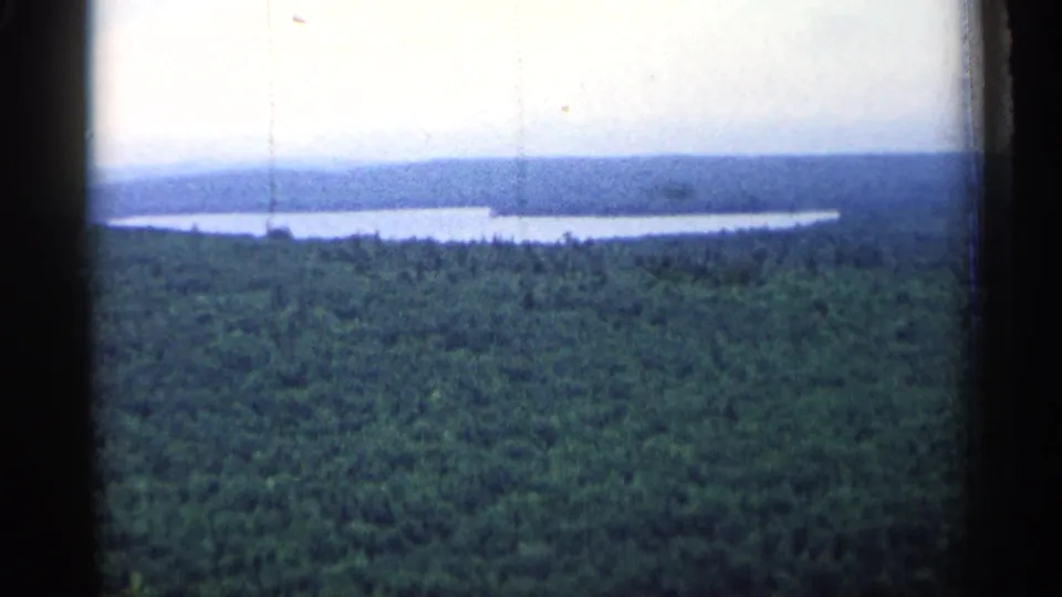 Vintage 8mm view of Michigan forest leading to a long lake, with hazy elevated terrain in the background.