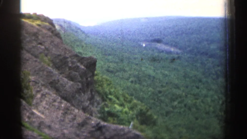 1960 Michigan 8mm frame from a rocky overlook above green forest, rolling hills, and distant water.