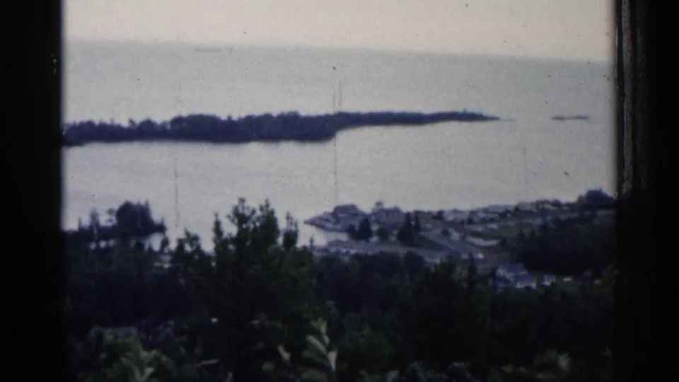 Elevated Copper Harbor area view shows foreground trees, small buildings, broad lake water, and a distant island or shoreline in soft 8mm focus.
