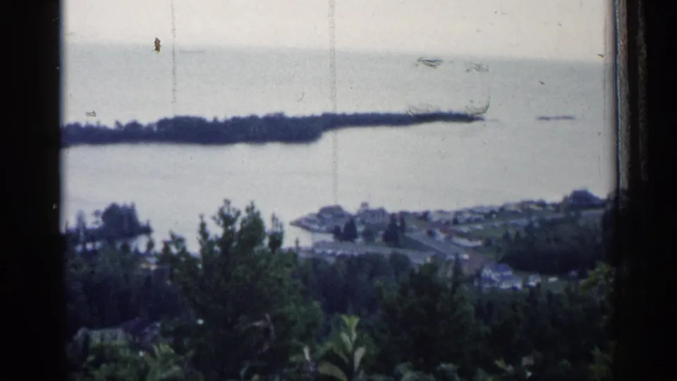 1960 Michigan 8mm view across a grey lake toward a small waterfront town and long wooded peninsula, framed by dark window edges.