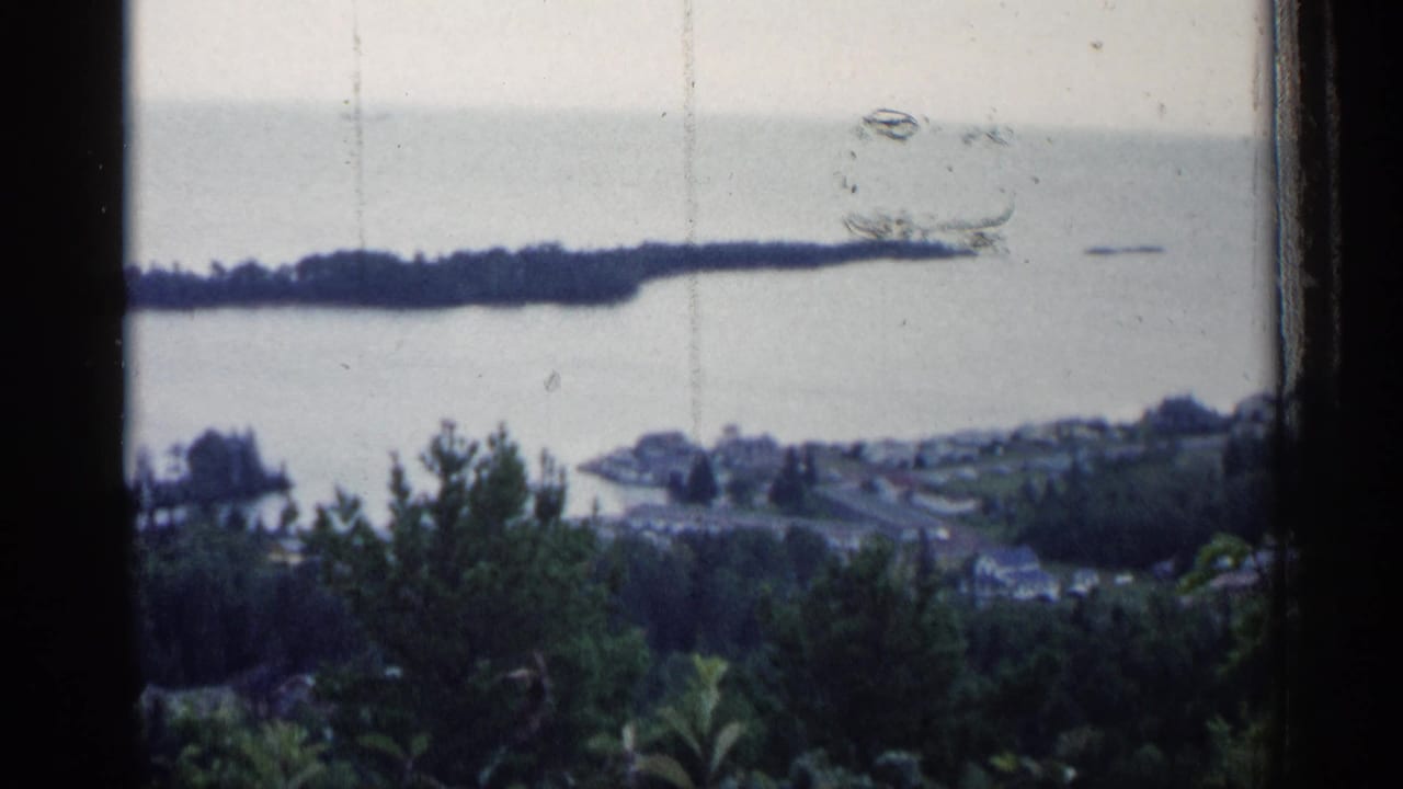 1960 Michigan home-movie frame showing a distant lakeside town, foreground greenery, and a wooded peninsula under overcast light.