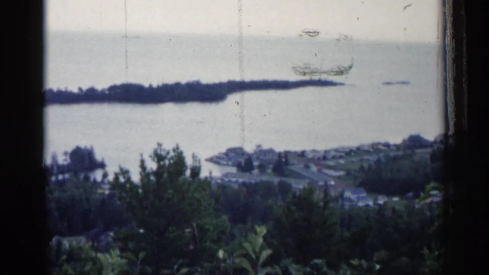 1960 Michigan 8mm frame of a muted lake overlook, with waterfront buildings below and a wooded strip extending across the water.