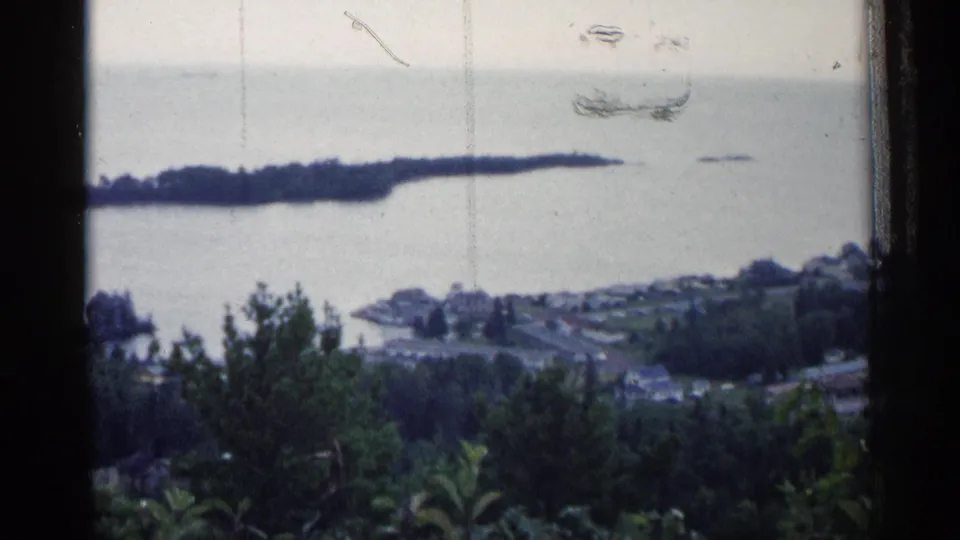 1960 Michigan lake overlook with a small waterfront community, wooded peninsula, foreground trees, and visible 8mm film scratches.