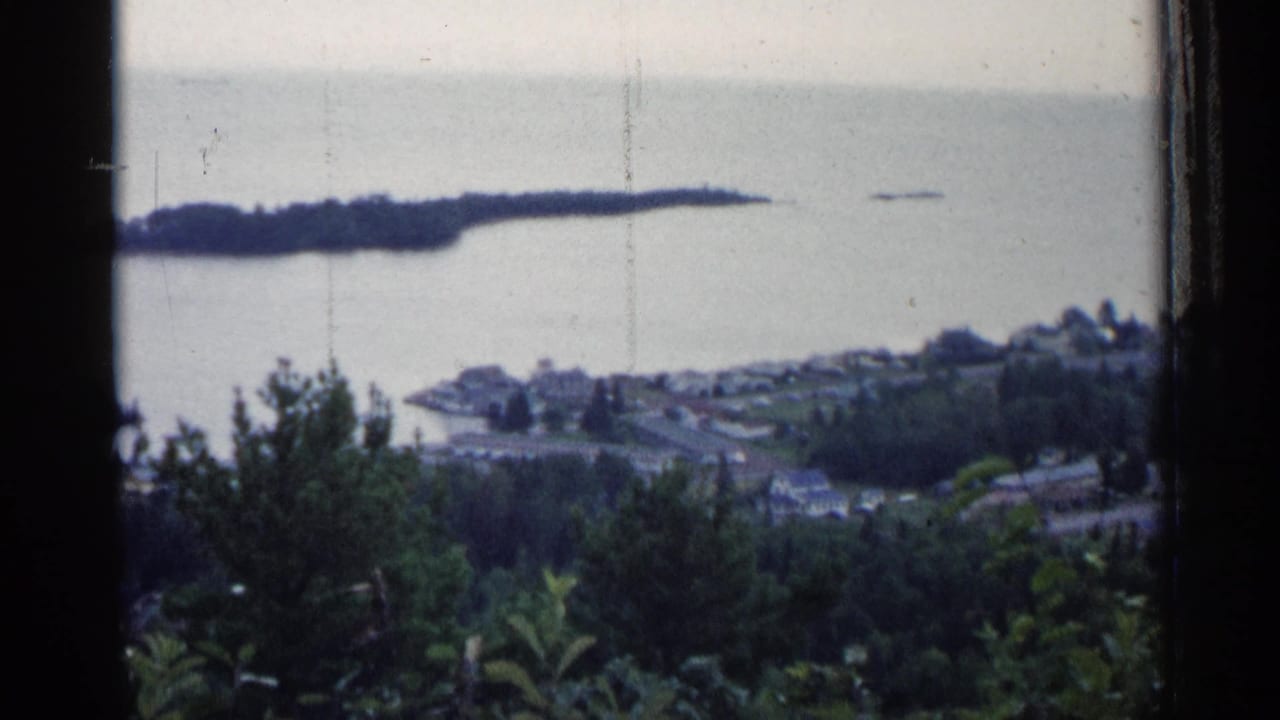 1960 Michigan home-movie view of a shoreline town, calm water, and a tree-covered island seen from an elevated window or overlook.