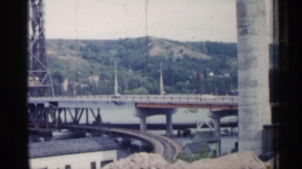1960 Michigan footage shows bridge traffic infrastructure over water, curving rail lines, and wooded hills beyond the shoreline.