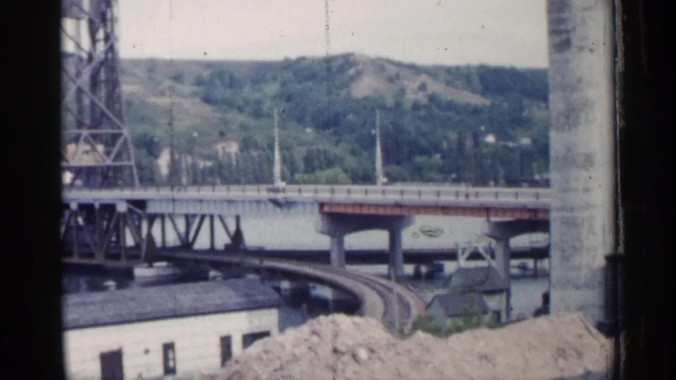 A 1960 Michigan home-movie frame looks past dark window edges toward a bridge, waterway, hillside, and rail curve below.