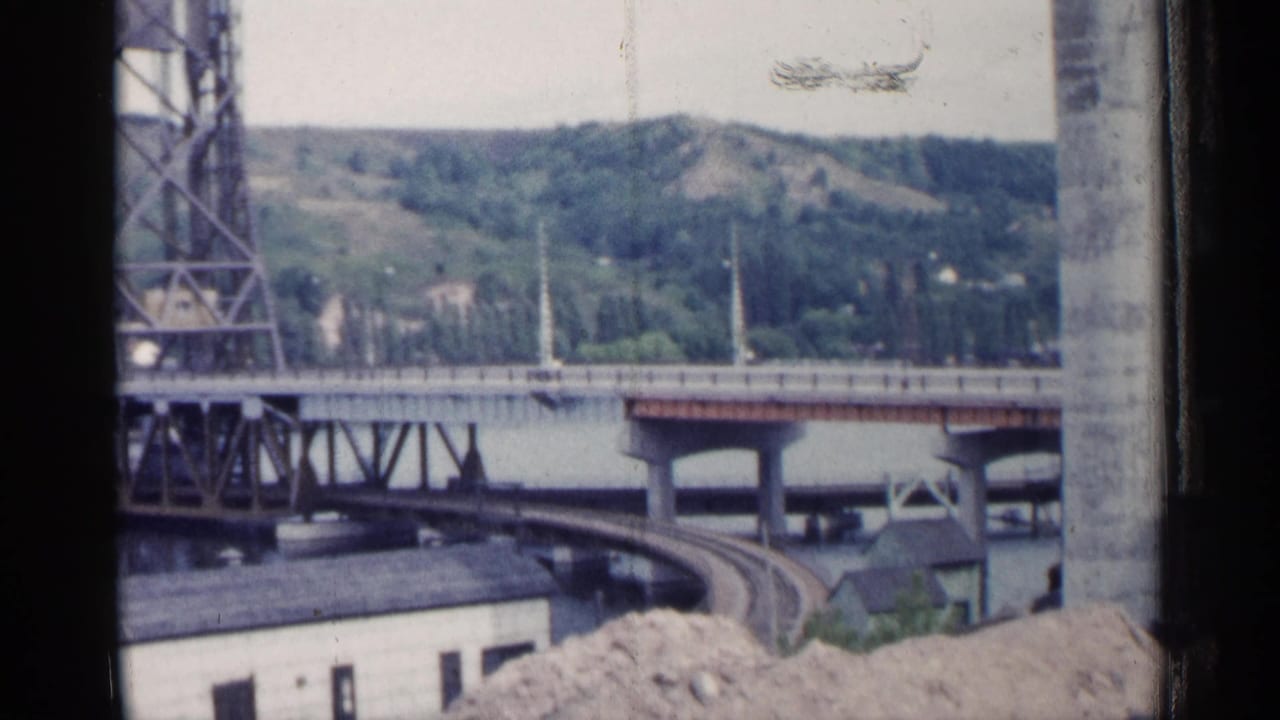 Steel truss bridge, river, curved rail line, buildings, and green hills seen through a window opening.