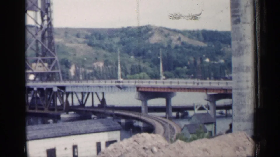 1960 Michigan 8mm view of a steel bridge by a river, with curved railway tracks, low buildings, and wooded hills beyond.