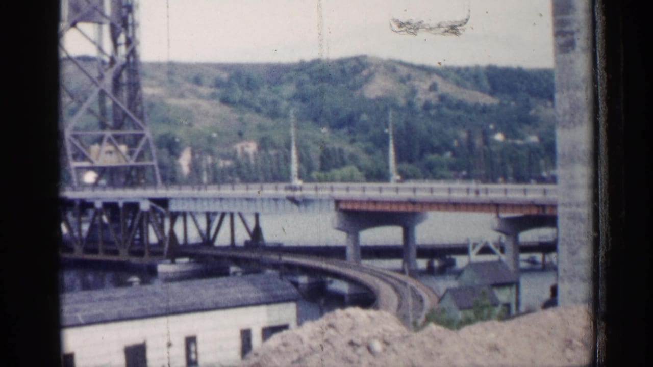 1960 Michigan home-movie frame showing bridge infrastructure over water, a curved rail line, and green hills across the river.