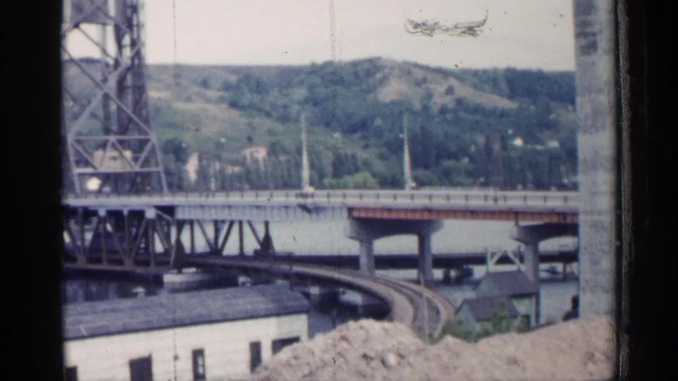 1960 Michigan home-movie frame showing bridge infrastructure over water, a curved rail line, and green hills across the river.