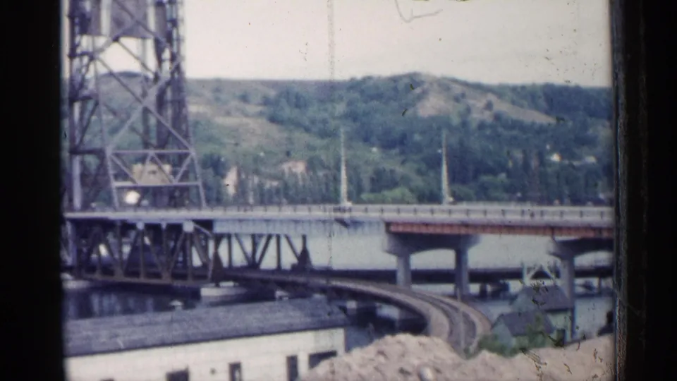 Soft 1960 Michigan 8mm frame of a steel bridge and curved railway line beside water, with wooded hills in the distance.