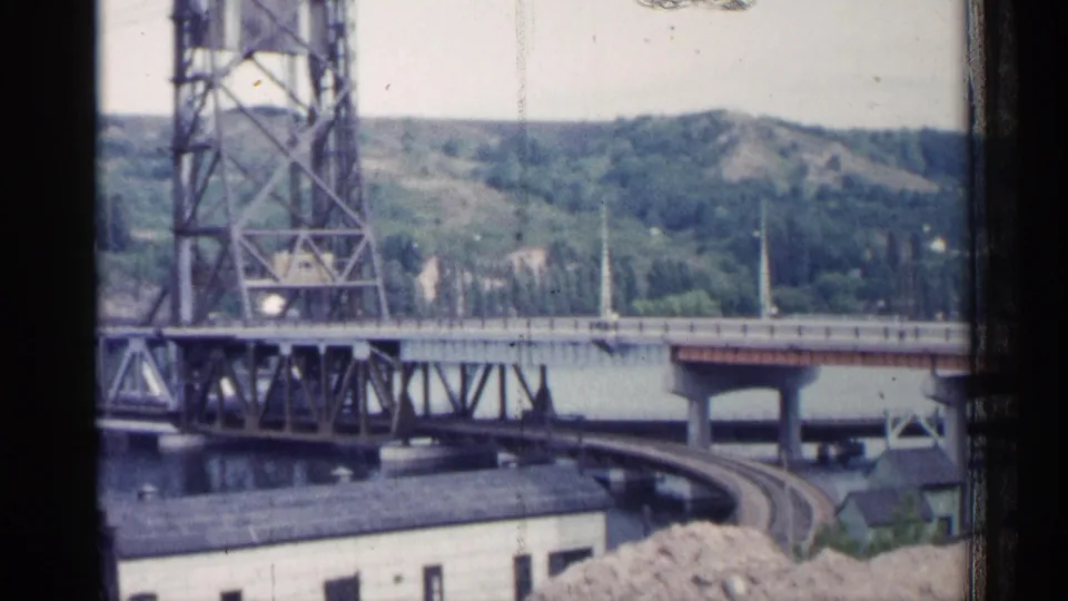 1960 Michigan 8mm view framed by dark edges, showing a steel bridge, river, rail curve, low buildings, and wooded hillside.
