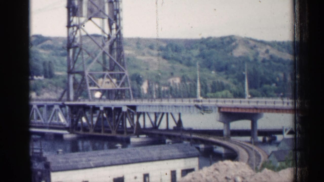 Steel lift bridge over a river with low buildings and wooded hills behind it.