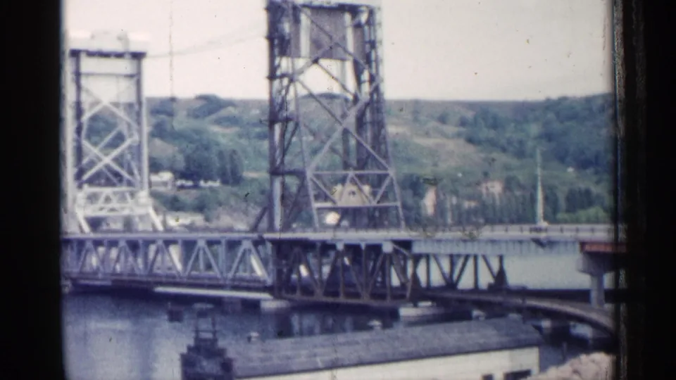 1960 Michigan 8mm frame showing the lift bridge structure raised over the river, with buildings, rail curve, and wooded hillside behind.
