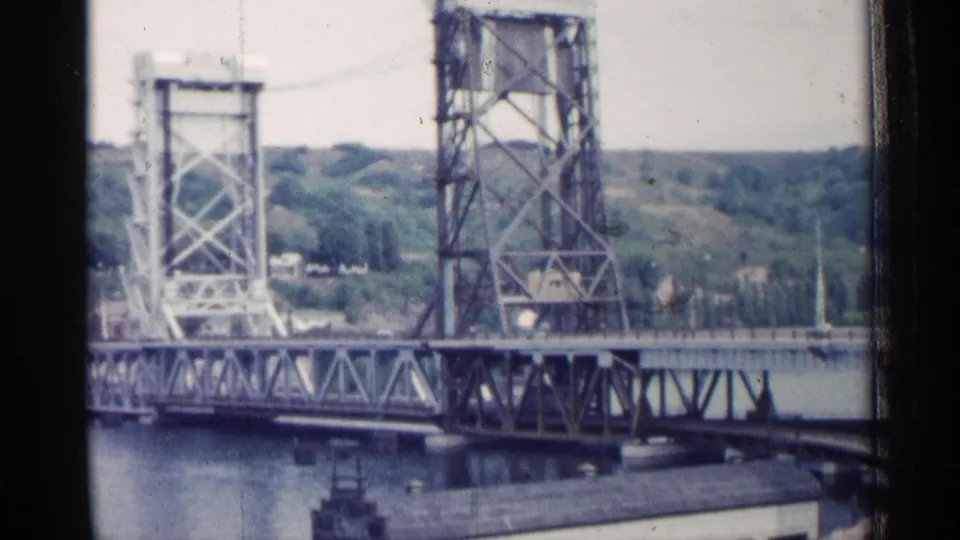 1960 Michigan home-movie frame of a steel lift bridge and raised tower over a river, with a low building and wooded hills beyond.