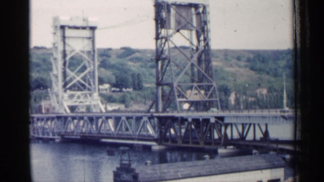 1960 Michigan 8mm frame showing a steel drawbridge with lift towers, truss span, water, low buildings, and trees beyond.