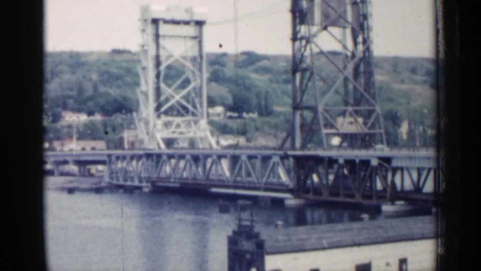 1960 Michigan 8mm still of a steel truss drawbridge crossing water, with lift towers, trees, and small buildings in the background.