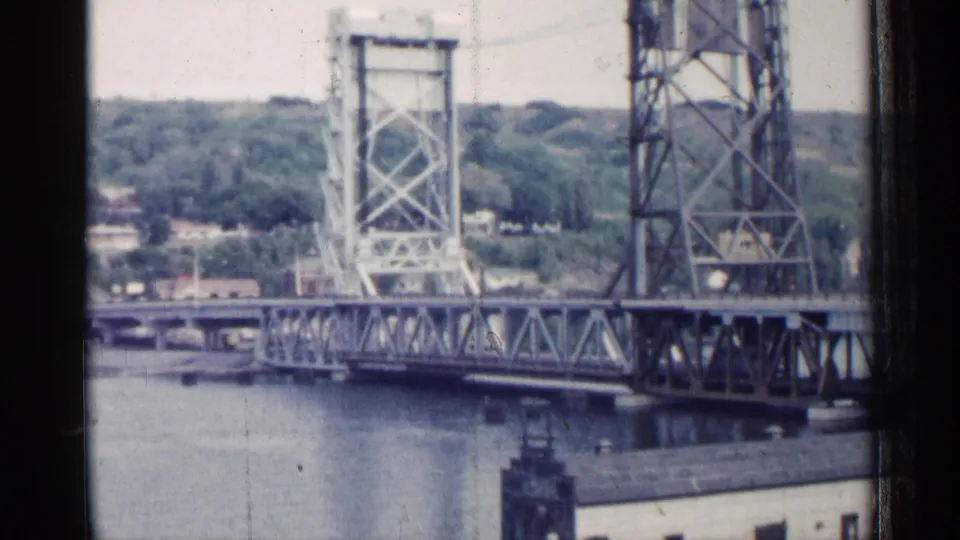 1960 Michigan frame showing the drawbridge structure, water below, distant wooded slope, and low waterfront buildings.