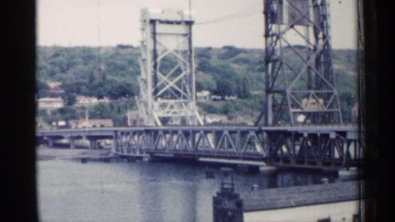 1960 Michigan home-movie frame showing a steel bascule bridge, river, and wooded residential hillside.