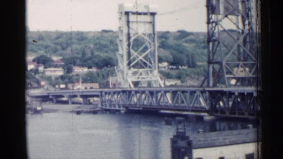 1960 Michigan bridge view with lift towers, steel trusswork, river, and distant hillside homes.
