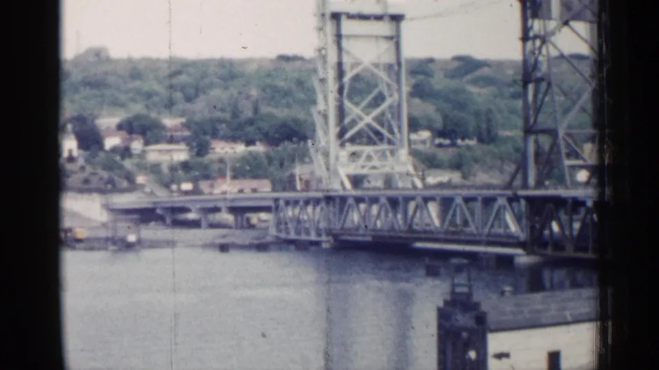 Soft 8mm frame of a Michigan bascule bridge over water, with hillside foliage and buildings beyond.
