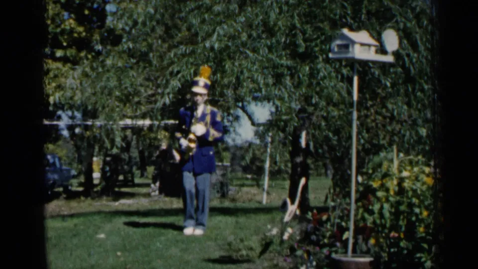 1960 Michigan home-movie frame of a uniformed saxophone player posed on a backyard lawn near garden plantings.