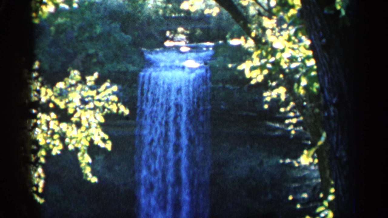 1960 Michigan 8mm frame showing a blue-toned waterfall framed by shaded tree trunks and sunlit forest foliage.