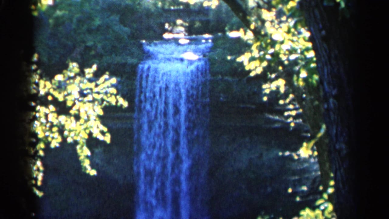 1960 Michigan 8mm frame with a blue waterfall partly framed by bright leaves, dark trunks, and shadowed woodland.