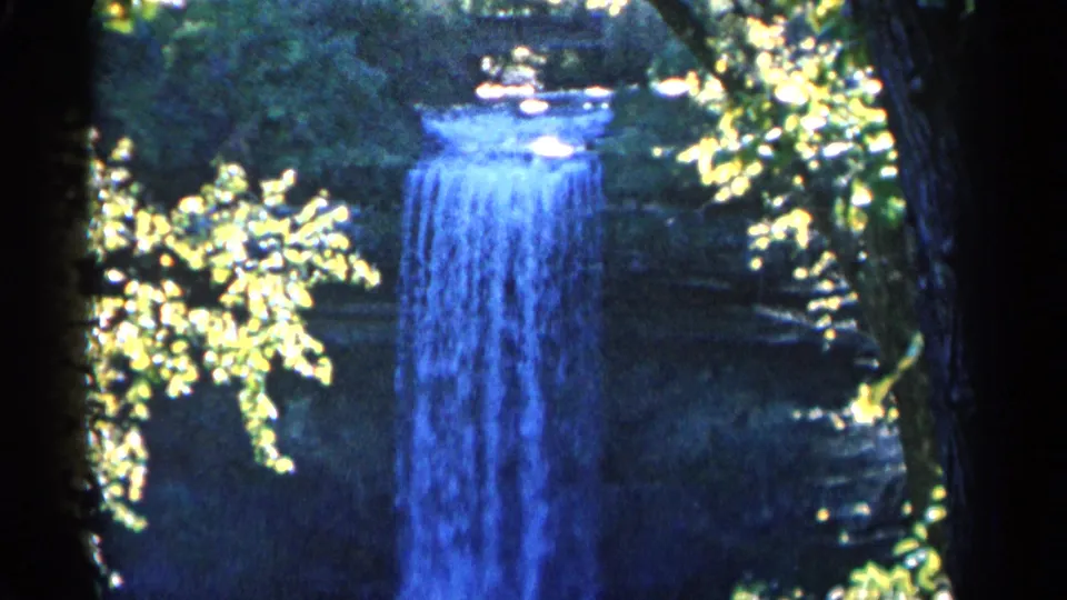 1960 Michigan 8mm frame of a forest waterfall framed by dark trunks, blue water, and yellow-green leaves in sunlight.