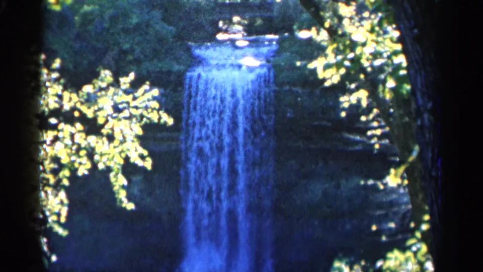 1960 Michigan 8mm frame showing a waterfall in a wooded setting, with bright leaves in the foreground and dark rock behind the cascade.