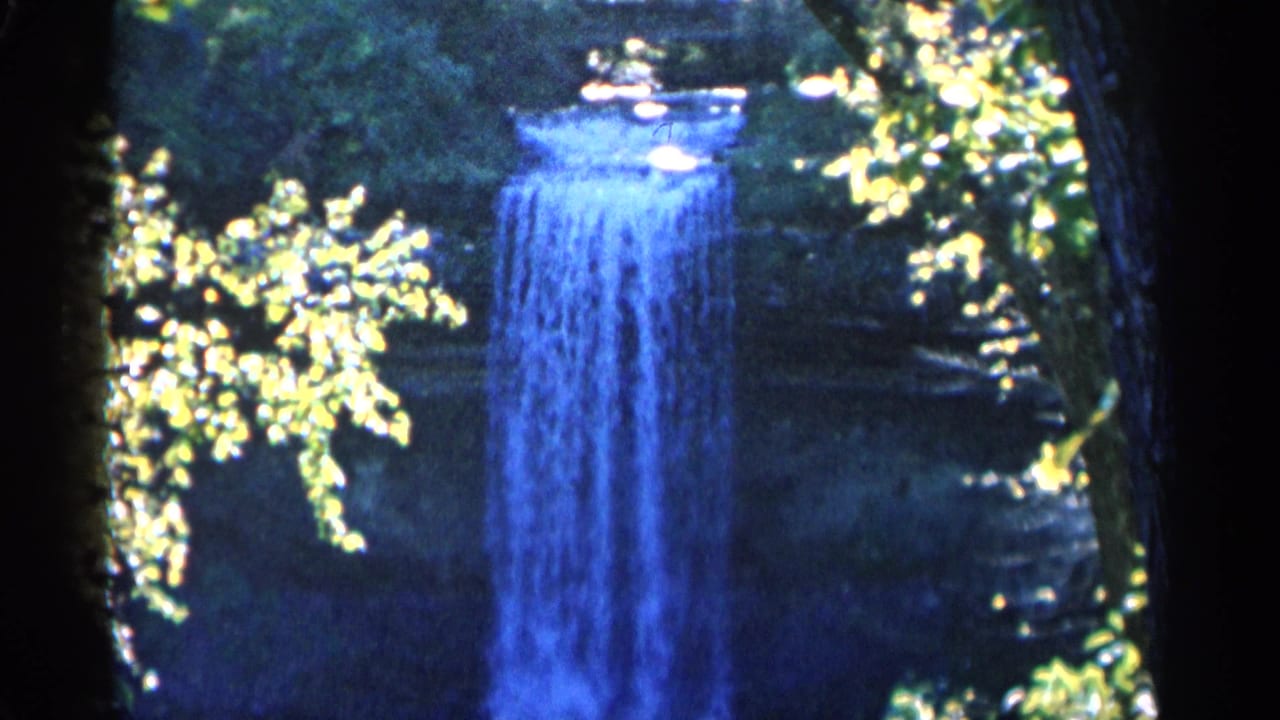 1960 Michigan 8mm frame with the waterfall held in view between trees, surrounded by shaded rock and bright summer foliage.
