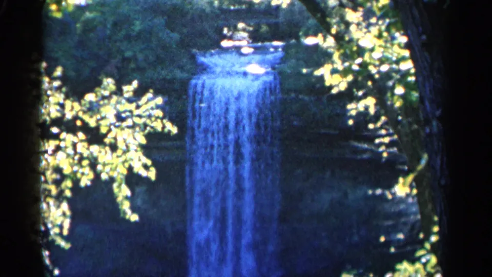 1960 Michigan 8mm frame of a blue waterfall framed by forest shadows, rough rock, tree trunks, and scattered sunlit leaves.