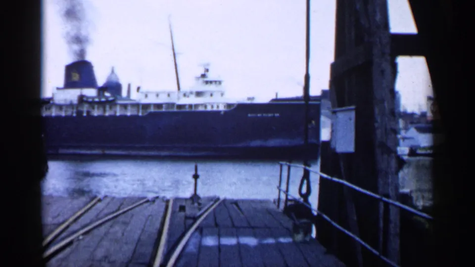 Ferry or lake vessel with smoke rising from its stack, seen beyond wooden planks and rail tracks on the pier.