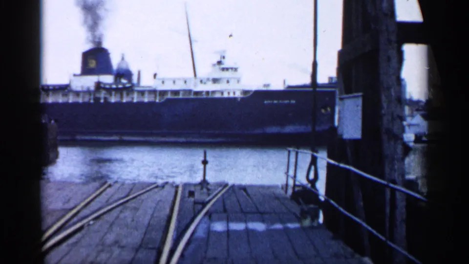 Large ferry along a waterfront, framed by dark pier supports with wooden decking and rails in the foreground.