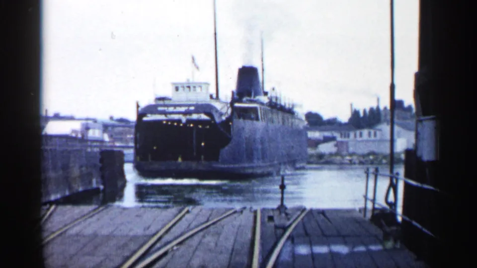 1960 Michigan harbor view of a dark-hulled ferry near the pier, with dock rails, water, smokestack, and shoreline structures visible.