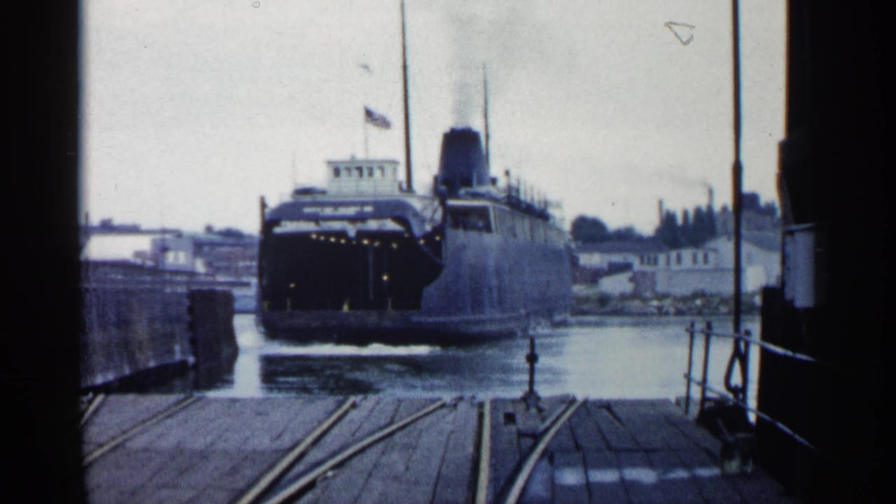 1960 Michigan home-movie frame of a large vessel approaching a dock, framed by pier structure with rail tracks leading toward the water.