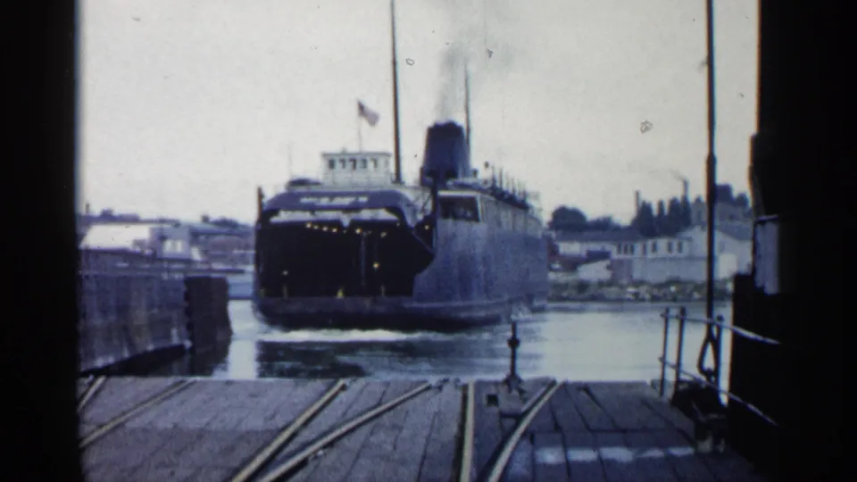 Large steamship in 1960 Michigan footage nearing a dock entrance, with rail tracks on wooden planks and industrial shoreline beyond.