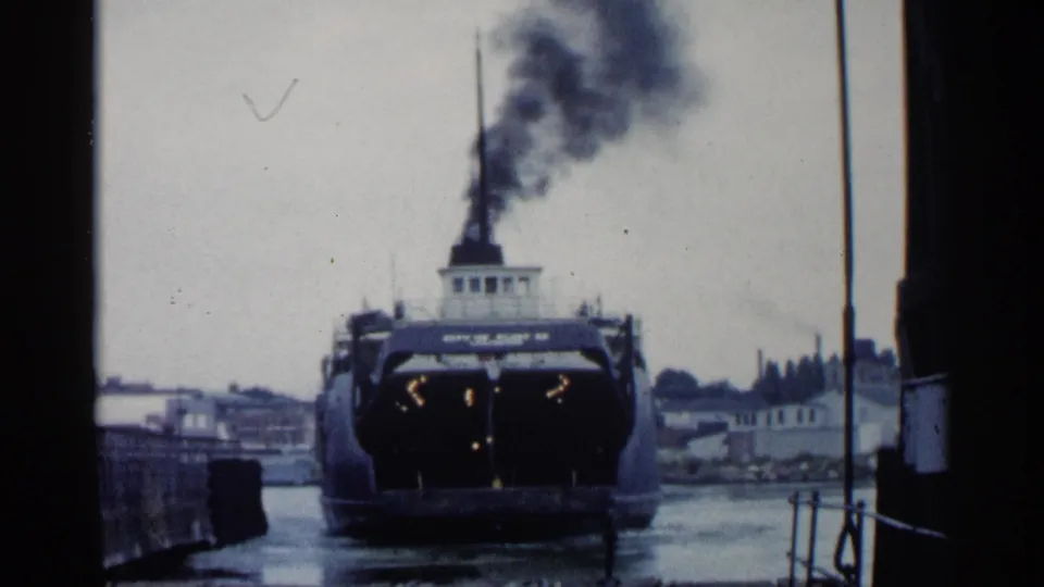1960 Michigan 8mm frame showing a ferry or steamship near a dock, with dark exhaust above the stack and waterfront buildings behind it.