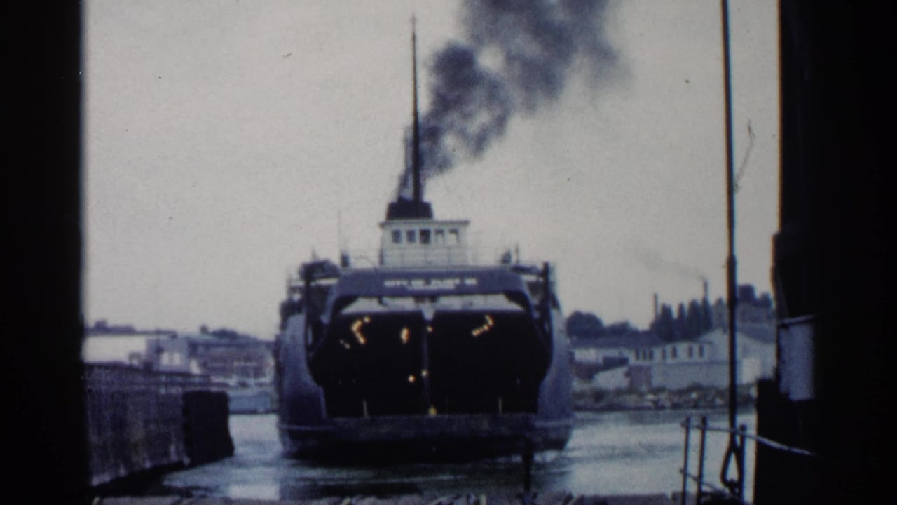 1960 Michigan 8mm frame with a rear view of a ferry or steamship at the dock, smoke drifting upward against a gray waterfront skyline.