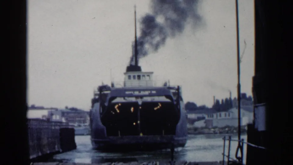 1960 Michigan 8mm frame with a rear view of a ferry or steamship at the dock, smoke drifting upward against a gray waterfront skyline.
