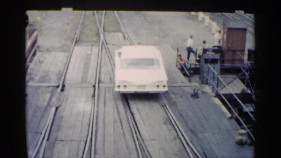 1960 Michigan 8mm frame showing a white mid-century American car moving along a wet wooden ferry deck with embedded rail tracks and distant people.