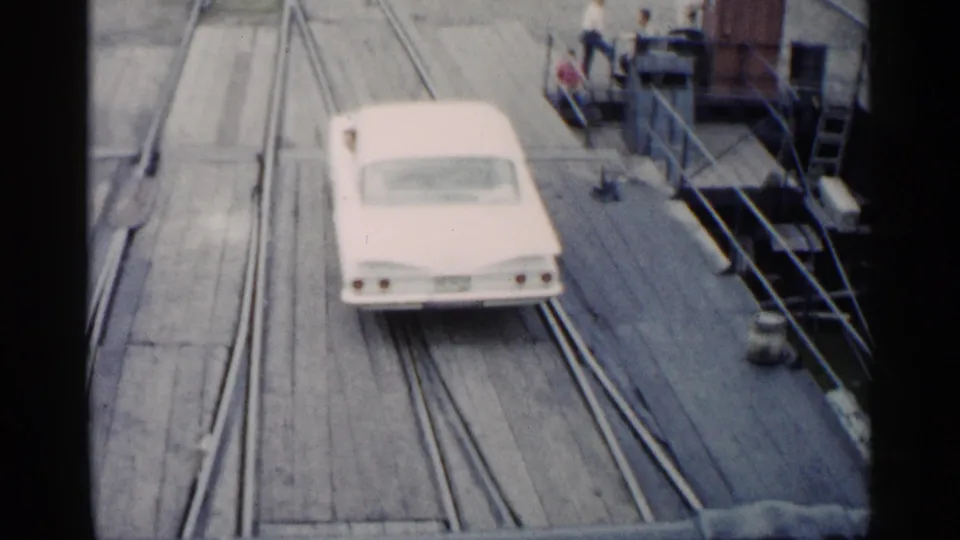 1960 Michigan 8mm frame with a white car driving on a ferry deck, rail tracks under it, and several distant people near dock structures.