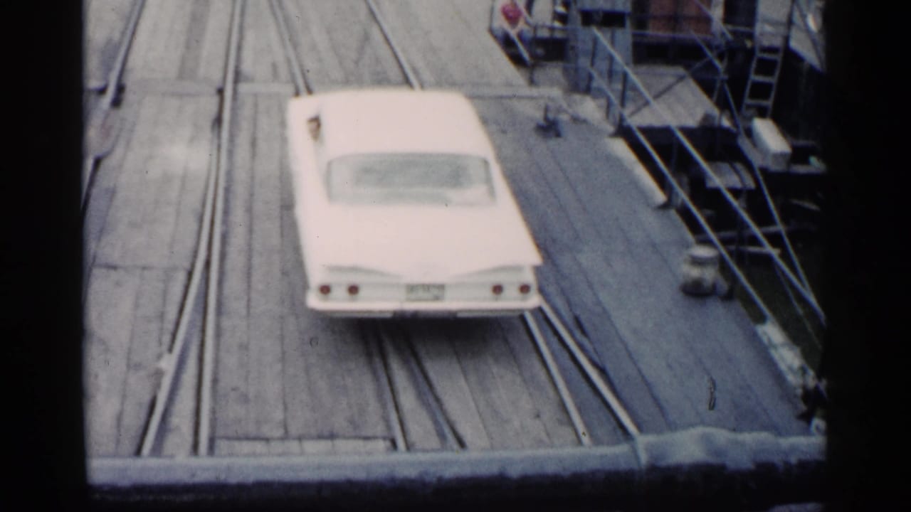 White classic car driving along a wooden ferry deck with rails and deck fittings nearby.