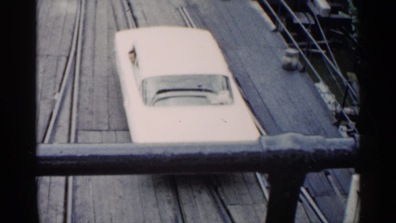 White car seen from above on a wooden ferry deck behind a foreground railing.