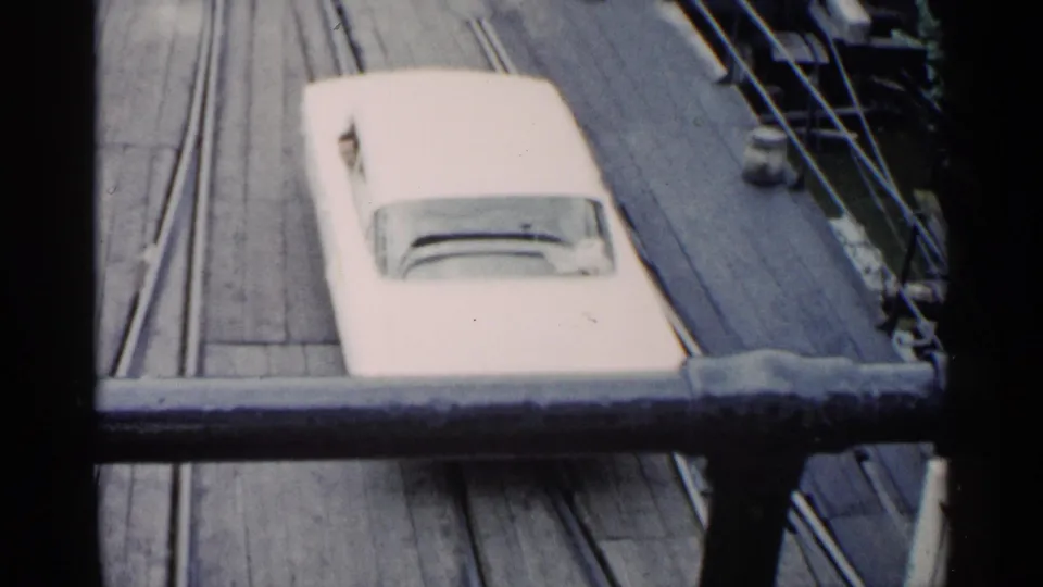 White mid-century car viewed from above on a ferry deck, partly blocked by a dark foreground rail in 1960 Michigan footage.