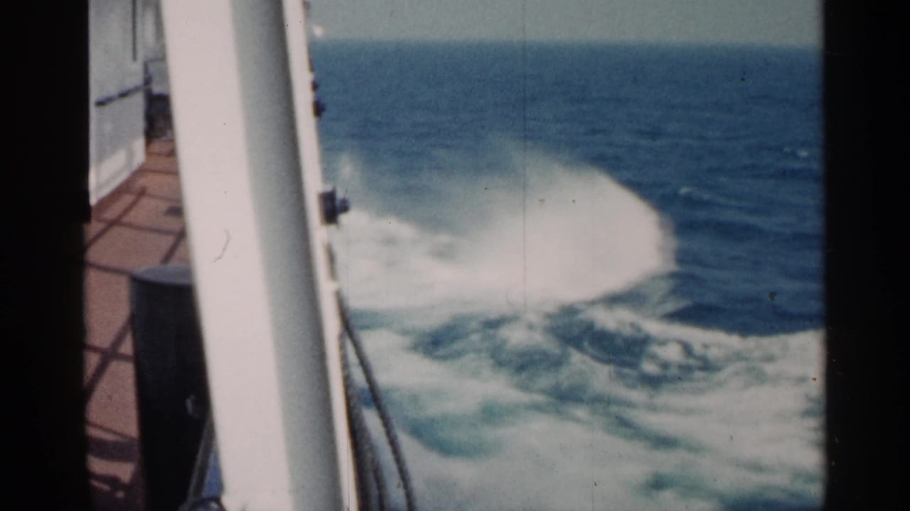 A 1960 8mm shipboard view shows white spray rising from the wake, with blue water and a pale horizon beyond.