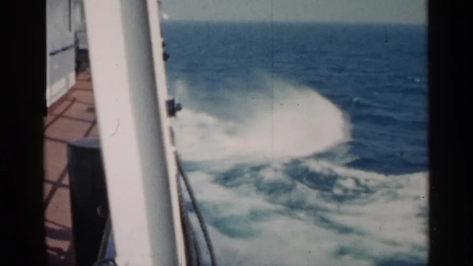 A 1960 8mm shipboard view shows white spray rising from the wake, with blue water and a pale horizon beyond.